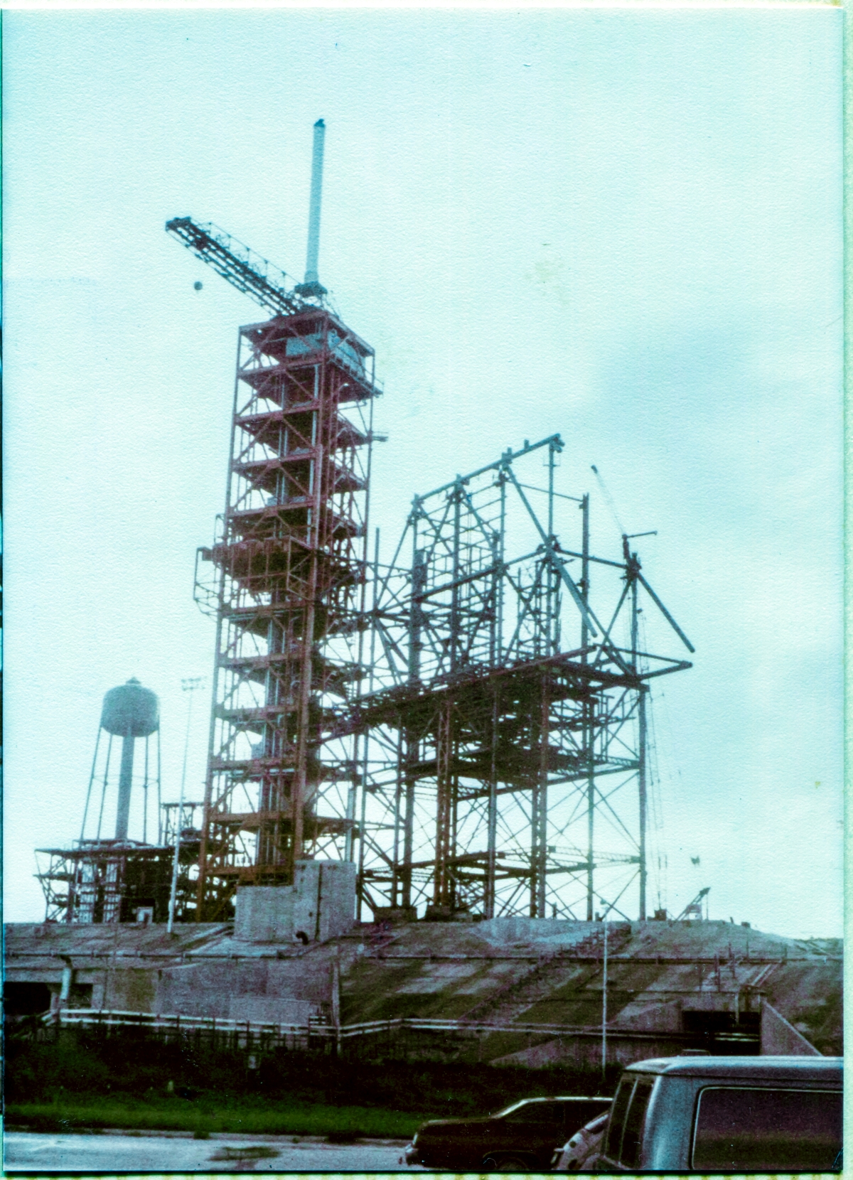 Image 003. Viewed from the parking lot just in front of the Sheffield Steel field trailer in the summer of 1980, the the incomplete steel skeleton of the Rotating Service Structure at Space Shuttle Launch Complex 39-B, Kennedy Space Center, Florida, stands supported on its temporary falsework support framing, growing into the sky, as it is assembled by the Union Ironworkers of Local 808, working for Wilhoit Steel Erectors. To its immediate left, stands the taller red framework of the Fixed Service Structure, topped by the Hammerhead Crane, and the Lightning Mast above that. Photo by James MacLaren.